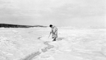 An Inuit woman with her baby on her back tries to hook a fish through a small tide crack, Coronation Gulf, Nunavut