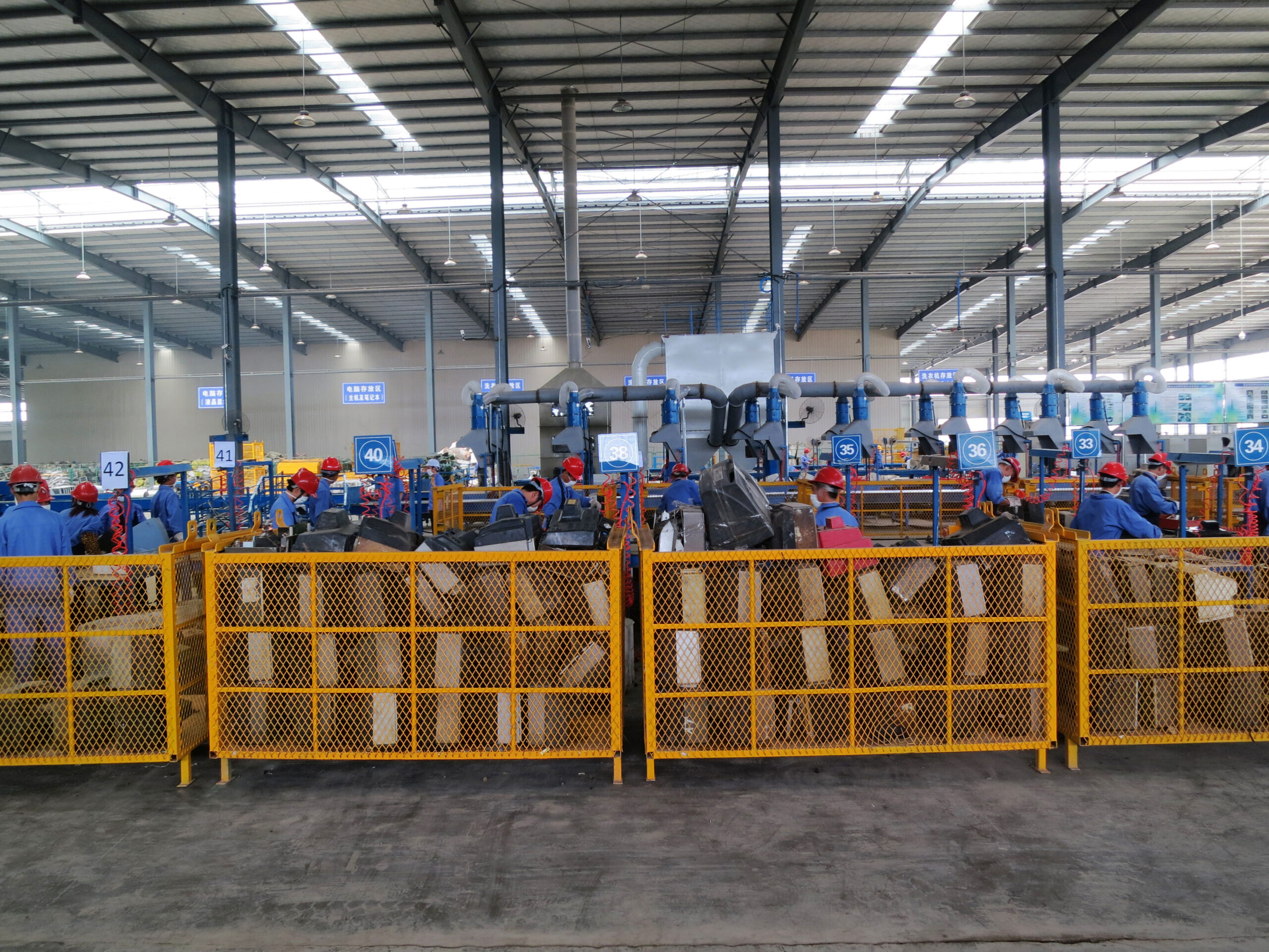 A row of yellow metal cage containers filled with e-waste being tended by people wearing blue suits and red hats.