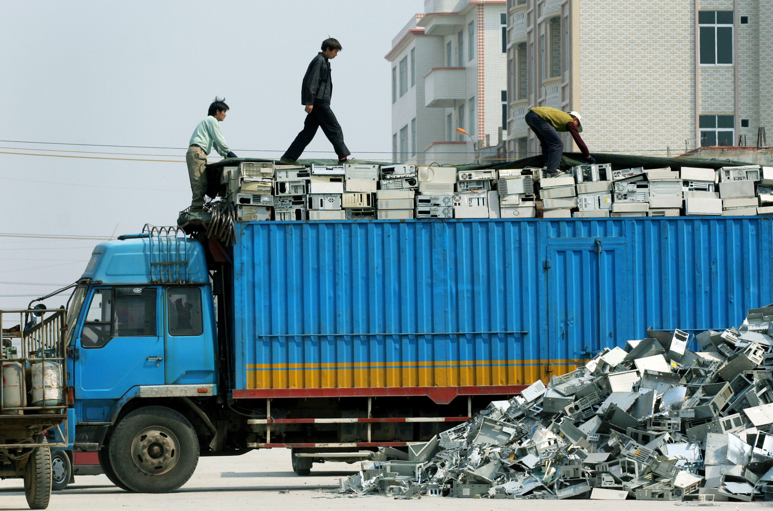 Three men standing on top of a blue truck loaded with computer waste next to a pile of more computer waste.
