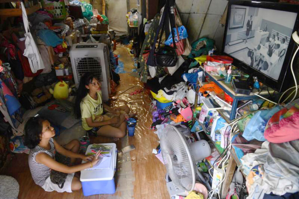 Two children sit on a cluttered floor during the COVID-19 pandemic, focused on a small TV broadcasting DLTV educational content