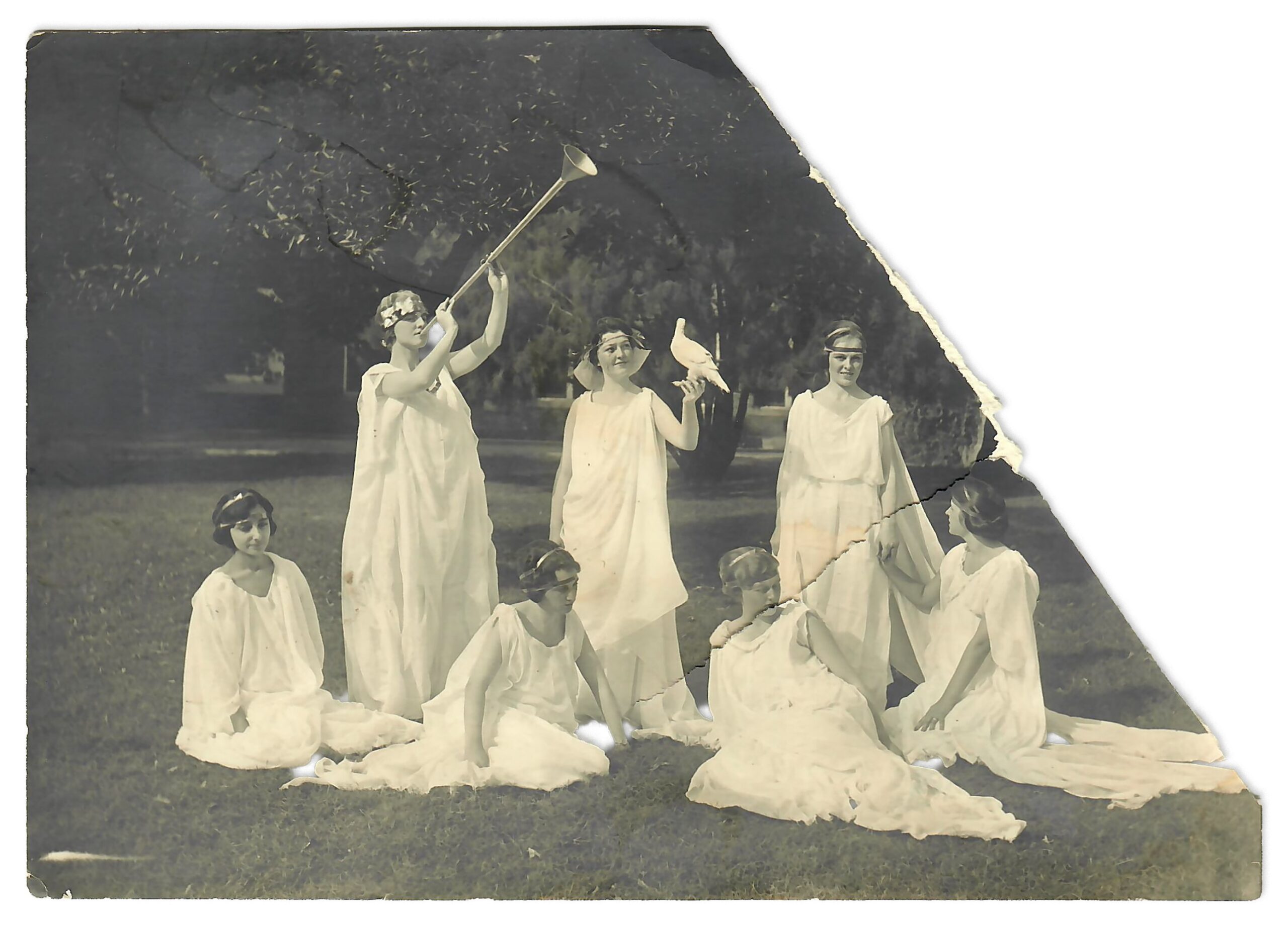 Black and white photo with one corner missing. Seven women in flowing white robes, one holds a dove, another plays a horn.