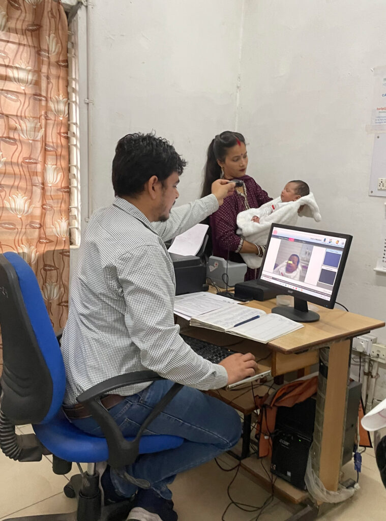 a woman holds a child, while a man sits behind a desk, looking at in image of the child through a camera held in his left hand